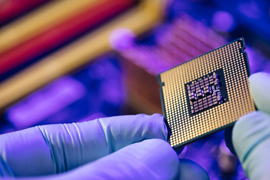 Close-up of blue-gloved hands holding a CPU, showing the gold contact pins, with colorful electronics blurred behind.